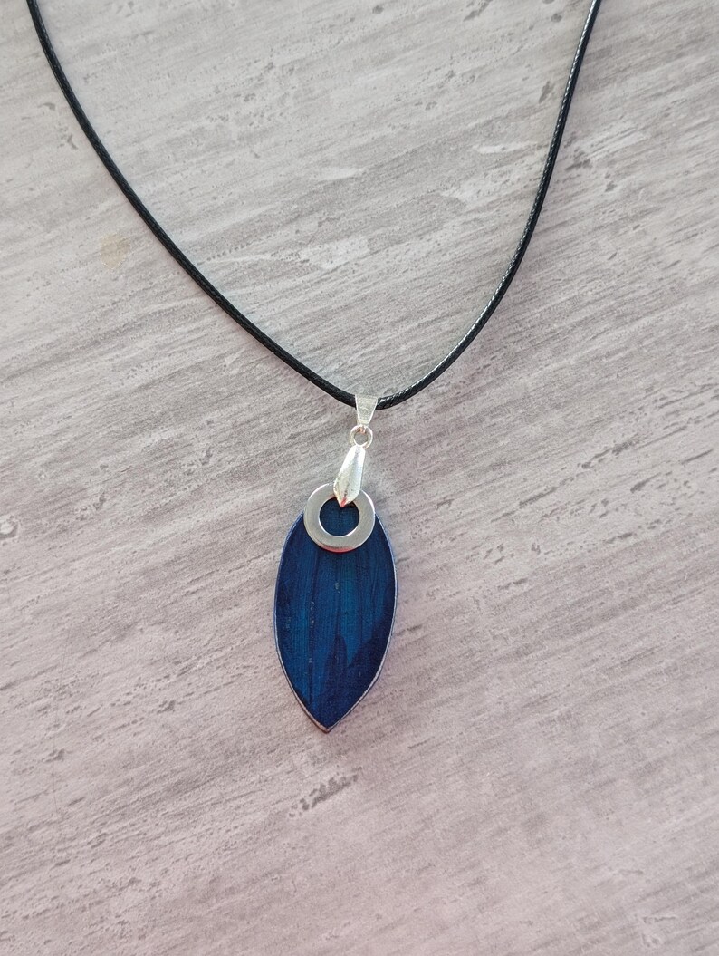 May include: A pendant necklace featuring a dark blue, leaf-shaped pendant with a silver-colored metal border and a silver-colored metal loop. The pendant hangs from a thin, black cord. The necklace is displayed on a light gray surface.