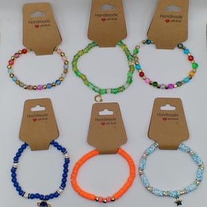 Op de afbeelding: Zes handgemaakte armbanden met verschillende gekleurde kralen en bedels. De armbanden liggen op bruine kaartjes met de tekst "Handmade with love".