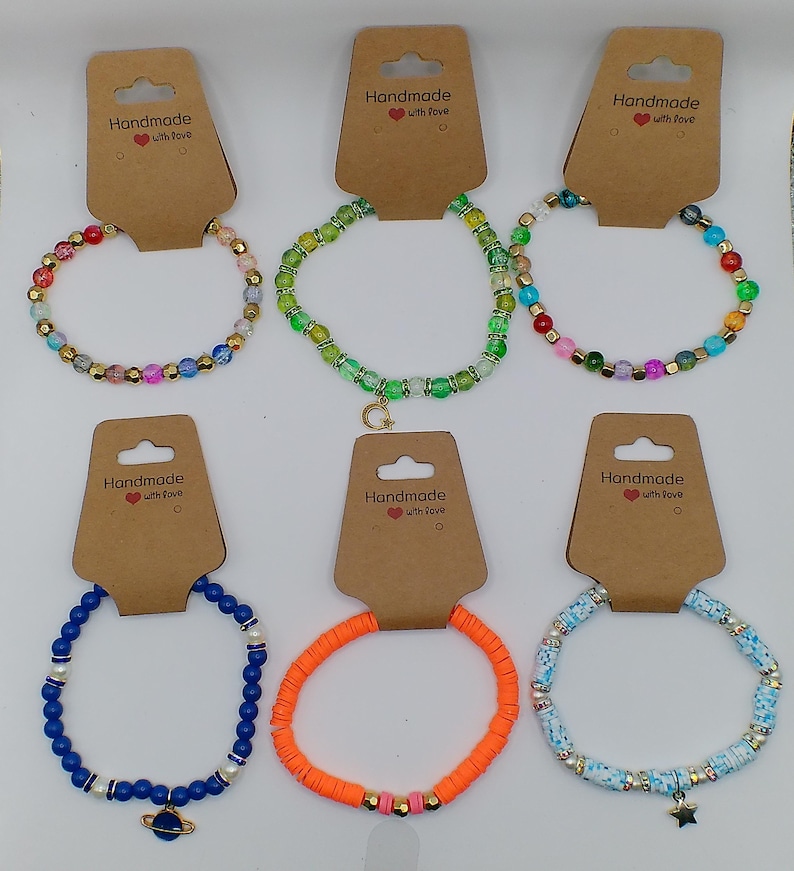 Op de afbeelding: Zes handgemaakte armbanden met verschillende gekleurde kralen en bedels. De armbanden liggen op bruine kaartjes met de tekst "Handmade with love".