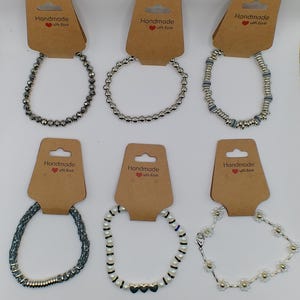 Op de afbeelding: Zes zilveren armbanden met verschillende ontwerpen, waaronder kralen, bedels en een hartbedel. De armbanden worden gepresenteerd op bruine kaartjes met de tekst "Handmade with love".