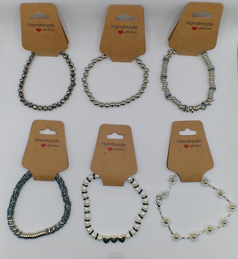 Op de afbeelding: Zes zilveren armbanden met verschillende ontwerpen, waaronder kralen, bedels en een hartbedel. De armbanden worden gepresenteerd op bruine kaartjes met de tekst "Handmade with love".