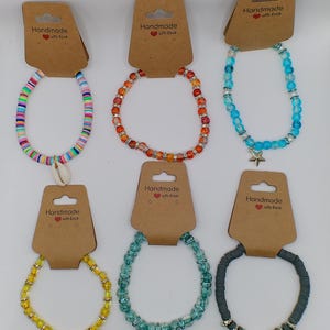 Op de afbeelding: Assortiment van acht handgemaakte kralenarmbanden in verschillende kleuren en stijlen. Sommige armbanden hebben een schelp- of sterbedel. Elke armband is verpakt op een bruine kaart met de tekst "Handmade with love".