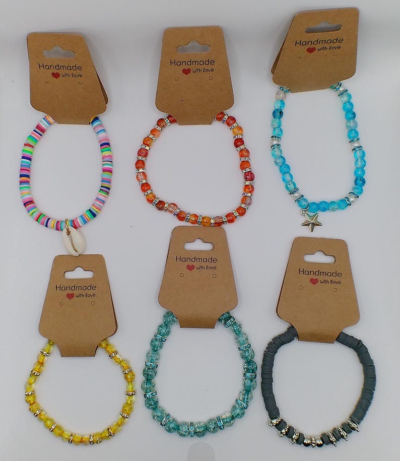 Op de afbeelding: Assortiment van acht handgemaakte kralenarmbanden in verschillende kleuren en stijlen. Sommige armbanden hebben een schelp- of sterbedel. Elke armband is verpakt op een bruine kaart met de tekst "Handmade with love".