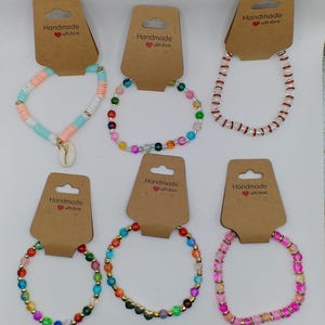 Op de afbeelding: Assortiment van kleurrijke kralenarmbanden met gouden accenten. Elke armband is handgemaakt en wordt geleverd met een bruin kaartje met de tekst "Handmade with love".
