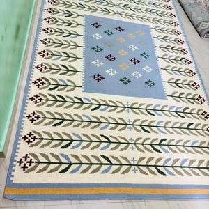 May include: A rectangular area rug featuring a cream-colored background with a repeating pattern of green and blue leaf-like branches. The center contains a light blue rectangular panel decorated with small, multi-colored cross-stitch motifs.