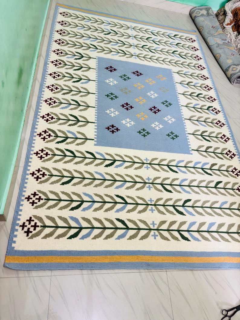 May include: A rectangular area rug featuring a cream-colored background with a repeating pattern of green and blue leaf-like branches. The center contains a light blue rectangular panel decorated with small, multi-colored cross-stitch motifs.