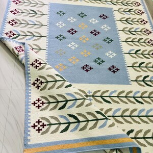 May include: A rectangular area rug featuring a cream-colored background with a repeating pattern of green and blue leaf-like stems. The center has a light blue rectangular panel decorated with small, multi-colored cross-stitch style floral motifs.