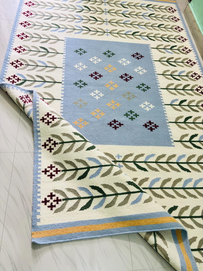 May include: A rectangular area rug featuring a cream-colored background with a repeating pattern of green and blue leaf-like stems. The center has a light blue rectangular panel decorated with small, multi-colored cross-stitch style floral motifs.