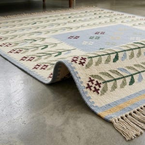 May include: A rectangular area rug featuring a cream-colored base with a repeating pattern of green leafy branches and small maroon cross-stitch flowers. The rug includes a light blue central panel, a yellow border, and off-white fringe at the ends.