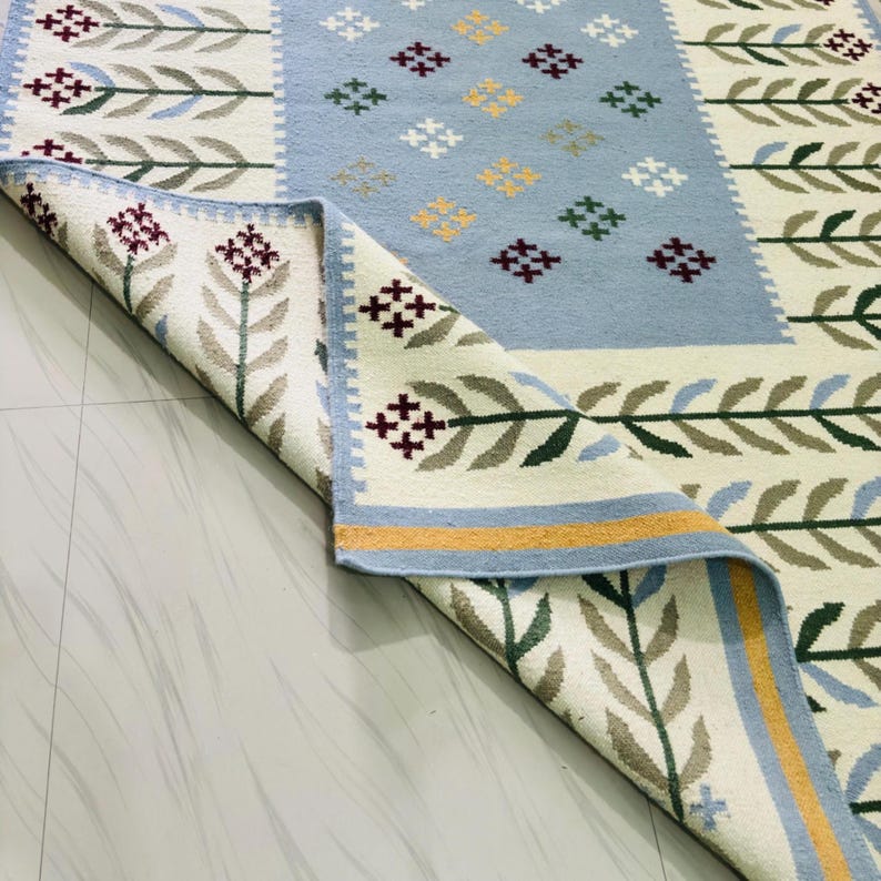 May include: A rectangular area rug featuring a cream-colored background with a repeating pattern of green and grey leaf stalks topped with maroon flowers. A central light blue panel displays a grid of small cross-shaped motifs in yellow, white, and maroon.