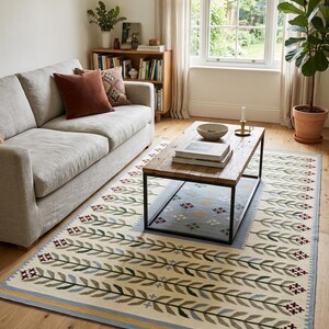 May include: A rectangular area rug featuring a cream-colored base with a repeating pattern of stylized green and blue leaf-like branches. The design includes small dark red cross-stitch accents and a border with light blue and yellow stripes.