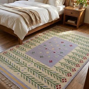 May include: A rectangular area rug featuring a cream-colored base with a central light purple rectangular panel decorated with small multi-colored floral motifs. The border displays repeating green leaf patterns and red flowers with fringe.