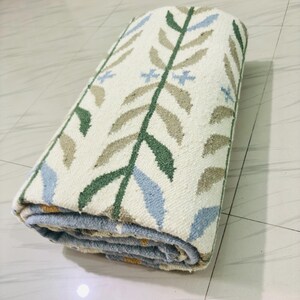 May include: A rolled area rug featuring an off-white base color with a repeating botanical pattern. The design consists of vertical stems in dark green with alternating light blue and tan leaves, accented by small light blue cross-shaped flowers.