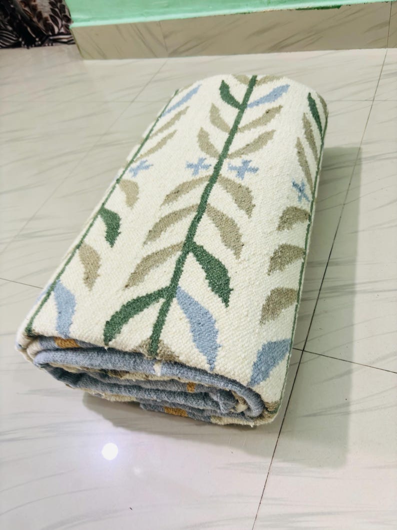 May include: A rolled area rug featuring an off-white base color with a repeating botanical pattern. The design consists of vertical stems in dark green with alternating light blue and tan leaves, accented by small light blue cross-shaped flowers.
