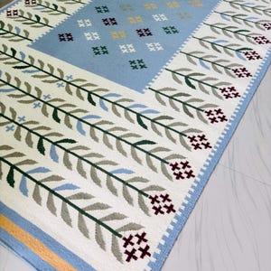 May include: A rectangular area rug featuring a central light blue panel decorated with small cross-stitched floral motifs in yellow, green, and dark red. The border displays repeating green and light blue leaf patterns on a white background.