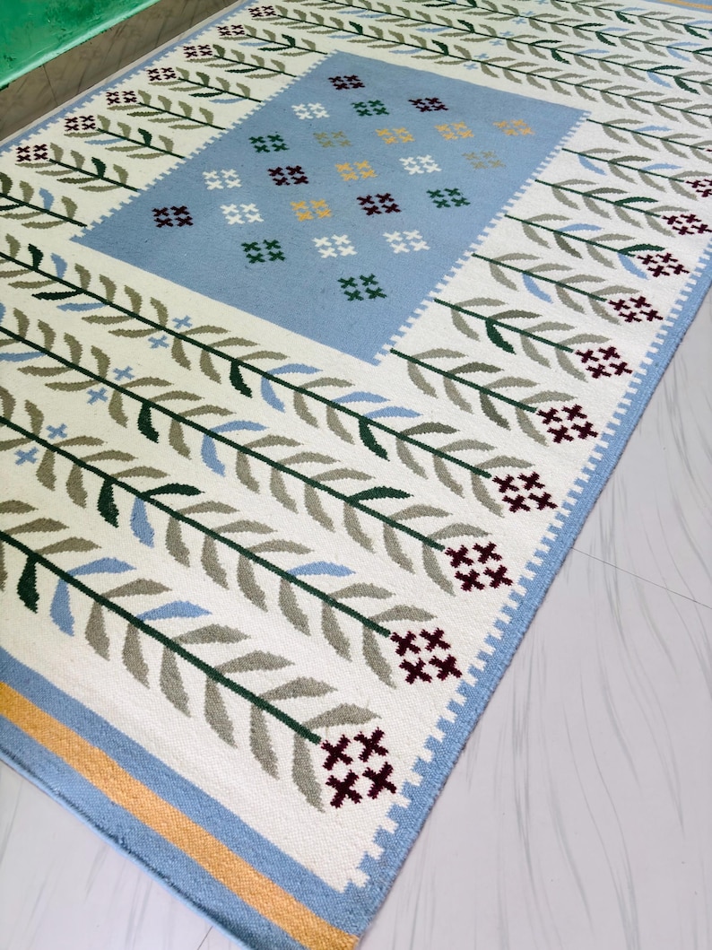 May include: A rectangular area rug featuring a central light blue panel decorated with small cross-stitched floral motifs in yellow, green, and dark red. The border displays repeating green and light blue leaf patterns on a white background.