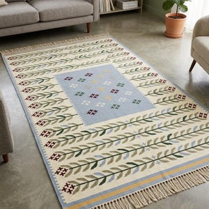 May include: A rectangular area rug featuring a cream-colored base with a central light blue panel decorated with small cross-stitch floral motifs. The border displays repeating green leaf vines and dark red flowers, finished with beige fringe.