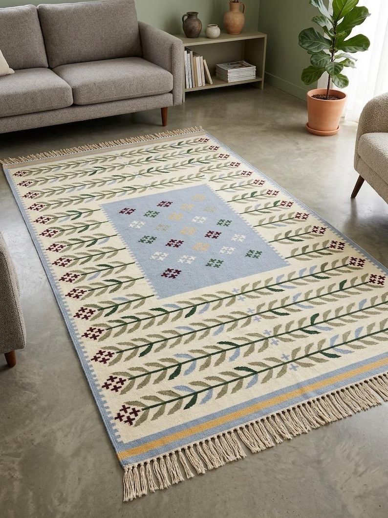 May include: A rectangular area rug featuring a cream-colored base with a central light blue panel decorated with small cross-stitch floral motifs. The border displays repeating green leaf vines and dark red flowers, finished with beige fringe.