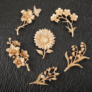 Pode incluir: Uma coleção de seis broches de metal em tom dourado com vários designs florais, incluindo um girassol, lírios do vale e flores de cerejeira. Algumas peças são acentuadas com pequenos cristais transparentes e pérolas artificiais.