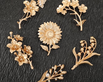 Può includere: Una collezione di sei spille floreali in metallo color oro. Il set include un girasole, un fiore con farfalla, mughetti e altre tre composizioni floreali distinte, alcune decorate con piccole perle bianche e cristalli trasparenti.