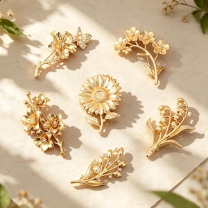Könnte beinhalten: Eine Sammlung von sechs goldfarbenen Metallbroschen mit verschiedenen Blumen- und Pflanzenmotiven, darunter eine Sonnenblume, ein Schmetterling auf einer Blume, Maiglöckchen und weitere Blüten, teilweise mit kleinen klaren Kristallen verziert.