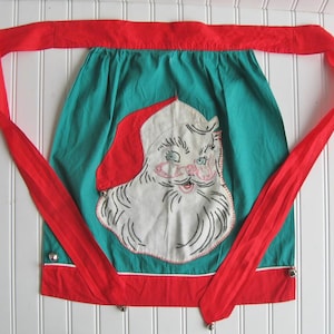 May include: A teal apron featuring a large white applique of a Santa Claus face with a red hat. The apron has a red waistband with long ties, a red bottom border with a thin white stripe, and small silver jingle bells attached to the edges.