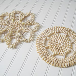 May include: Two decorative seashell coasters. One is a starburst shape, and the other is round. Both are made of small, cream-colored seashells and are placed on a white, slatted surface.