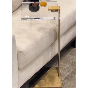 May include: A modern side table with a clear acrylic top and a gold-colored metal frame. The table is positioned next to a cream-colored sofa. A small black bowl and a glass of orange liquid sit on the table.