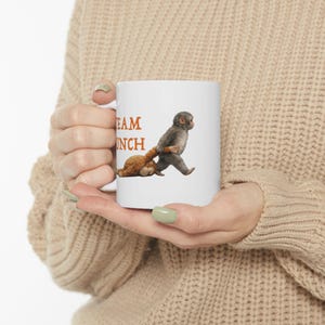 May include: White ceramic mug featuring a cartoon illustration of a monkey pulling a teddy bear. The mug has the words "TEAM LUNCH" printed in orange. The mug is held by a person.
