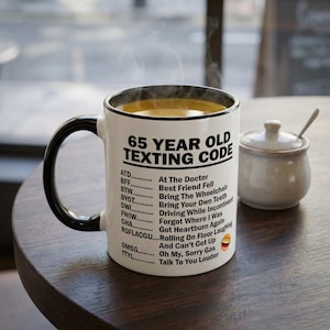Op de afbeelding: Een witte keramische mok met een zwarte handgreep en rand, gevuld met een warme drank. De mok heeft de tekst "65 YEAR OLD TEXTING CODE" en een lijst met acroniemen met hun betekenissen. Een kleine suikerpot en lepel staan op de tafel.