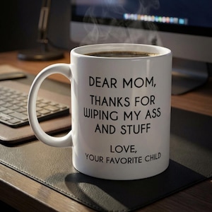 May include: White ceramic mug filled with dark liquid, possibly coffee, with steam rising. The mug has a curved handle and text that reads "DEAR MOM, THANKS FOR WIPING MY ASS AND STUFF, LOVE, YOUR FAVORITE CHILD."