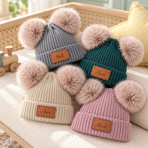 May include: Four ribbed knit beanies in gray, dark green, cream, and dusty pink. Each hat features two large, fluffy, light brown faux fur pom-poms and a rectangular brown leather patch with the names Hale, Pearl, Joyce, and Rock.