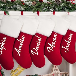 Puede incluir: Seis calcetines navideños rojos con puños de piel sintética blanca cuelgan de una repisa. Cada calcetín está personalizado con un nombre en escritura blanca: Sophia, Amelia, Daniel, Mason y Jackson. La decoración festiva rodea los calcetines.