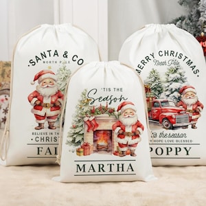 May include: Three white Christmas gift bags with drawstring closures, each adorned with a Santa Claus design. The bags display festive text such as "Santa & Co," "Merry Christmas," and "'Tis the Season." The illustrations include a fireplace and a red truck.