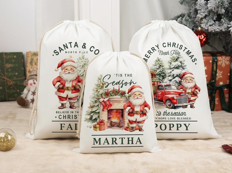 May include: Three white Christmas gift bags with drawstring closures, each adorned with a Santa Claus design. The bags display festive text such as "Santa & Co," "Merry Christmas," and "'Tis the Season." The illustrations include a fireplace and a red truck.