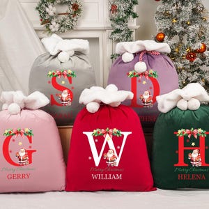 May include: A selection of Christmas gift bags in shades of pink, red, green, grey, and purple. Each bag displays a large initial, a Santa Claus illustration, and the words "Merry Christmas" plus a name. The bags are trimmed with white fluffy material.