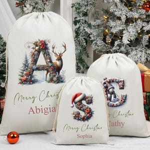 May include: Three cream-coloured drawstring Christmas gift bags with festive designs. The largest bag displays a floral "A" with a deer, the middle bag has a floral "S" with a Santa hat, and the smallest bag has a floral "C" with a snowman. Each bag has a name and "Merry Christmas" text.