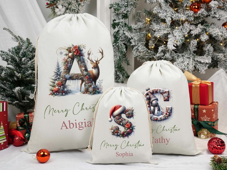May include: Three cream-coloured drawstring Christmas gift bags with festive designs. The largest bag displays a floral "A" with a deer, the middle bag has a floral "S" with a Santa hat, and the smallest bag has a floral "C" with a snowman. Each bag has a name and "Merry Christmas" text.