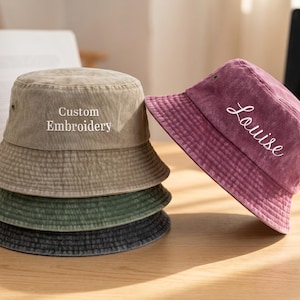 May include: A stack of four corduroy bucket hats in shades of tan, green, and charcoal gray. One burgundy hat is angled to the side, embroidered with the name "Louise". The top hat reads "Custom Embroidery".