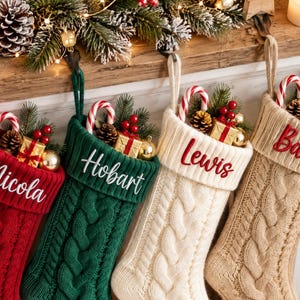 Op de afbeelding: Vier kerstsokken van kabelgebreid materiaal in rood, groen, crème en beige, elk met een omgeslagen boord geborduurd met namen zoals Nicola, Hobart en Lewis. De sokken zijn gevuld met zuurstokken, dennentakjes en kleine cadeautjes.