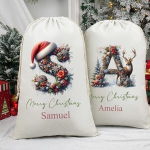 May include: Two cream-coloured Christmas gift bags with drawstring closures. One bag features a floral "S" topped with a Santa hat, the other a floral "A" with a deer. Both bags have "Merry Christmas" and a name printed on them.