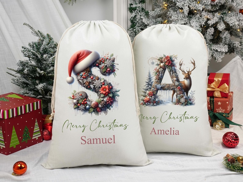 May include: Two cream-coloured Christmas gift bags with drawstring closures. One bag features a floral "S" topped with a Santa hat, the other a floral "A" with a deer. Both bags have "Merry Christmas" and a name printed on them.