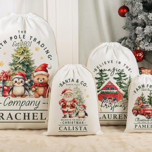 May include: Four white Christmas gift bags with drawstring closures. Each bag features a unique festive design, including Santa Claus, a penguin, a red truck, and a Christmas tree. The bags are personalised with names.