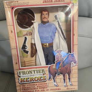 Puede incluir: Una figura de acci&oacute;n de Jesse James "Frontier Heroes" en caja. La figura lleva una camisa azul, vaqueros y un abrigo blanco. La caja incluye un rifle, una pistola y un sombrero de vaquero. La caja tambi&eacute;n presenta un dibujo de un vaquero a caballo.