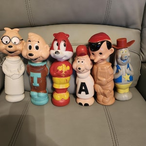 May include: A collection of six vintage plastic character figures. The set includes figures with distinct features such as glasses, a letter A, a red cap, and a cowboy hat. These molded toys display various colors including white, brown, red, blue, and yellow.