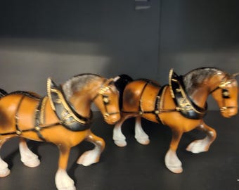 May include: Two brown draft horse figurines with white lower legs and hooves. Each horse wears a black harness with gold-colored trim and detailing. The horses are depicted in a walking pose with one front leg raised, featuring textured manes.