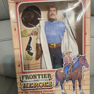 Puede incluir: Una figura de acci&oacute;n de Jesse James de Frontier Heroes en caja. La figura viste camisa azul, pantalones vaqueros, abrigo blanco y hebilla dorada. La caja presenta un dibujo de un vaquero a caballo e incluye el texto "12" posable figure".
