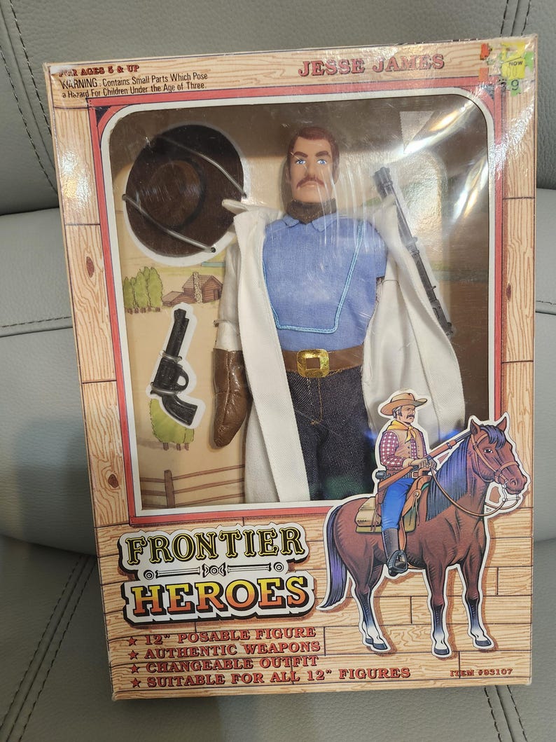 Puede incluir: Una figura de acci&oacute;n de Jesse James de Frontier Heroes en caja. La figura viste camisa azul, pantalones vaqueros, abrigo blanco y hebilla dorada. La caja presenta un dibujo de un vaquero a caballo e incluye el texto "12" posable figure".