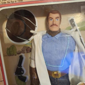 Puede incluir: Una figura de acci&oacute;n de Jesse James en su embalaje original. La figura viste una camisa azul, vaqueros y un abrigo blanco. Tiene un bigote marr&oacute;n y un sombrero marr&oacute;n. El embalaje incluye el texto "Small Parts Which Pose Under the Age of Three."