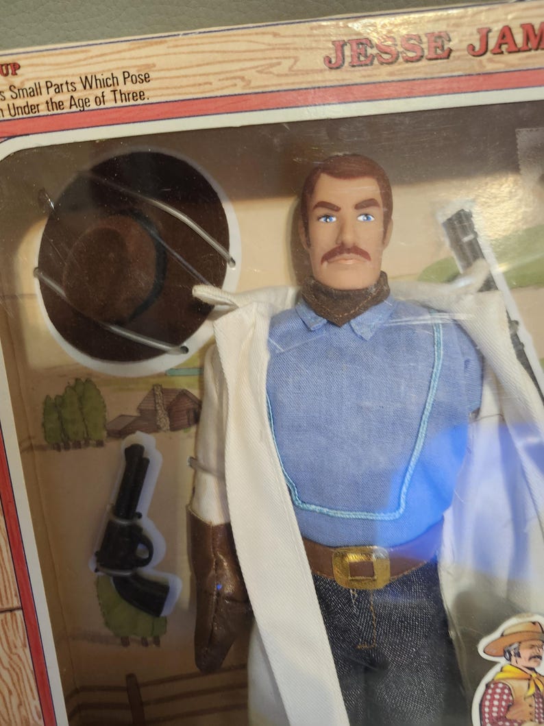 Puede incluir: Una figura de acci&oacute;n de Jesse James en su embalaje original. La figura viste una camisa azul, vaqueros y un abrigo blanco. Tiene un bigote marr&oacute;n y un sombrero marr&oacute;n. El embalaje incluye el texto "Small Parts Which Pose Under the Age of Three."